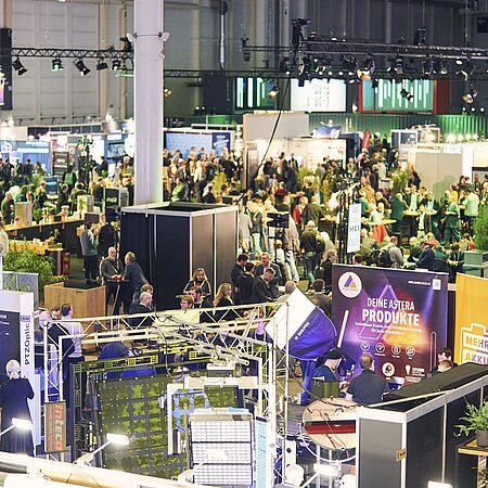 ebhafte Messehalle mit zahlreichen Ständen und Besuchern. Im Vordergrund sind Ausstellungsstände mit technischen Geräten und der Aufschrift „Deine Astera Produkte“ zu sehen. Pflanzen dienen als Dekoration, im Hintergrund sind weitere Stände und viele Menschen erkennbar.