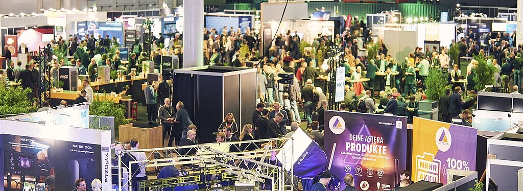 ebhafte Messehalle mit zahlreichen Ständen und Besuchern. Im Vordergrund sind Ausstellungsstände mit technischen Geräten und der Aufschrift „Deine Astera Produkte“ zu sehen. Pflanzen dienen als Dekoration, im Hintergrund sind weitere Stände und viele Menschen erkennbar.
