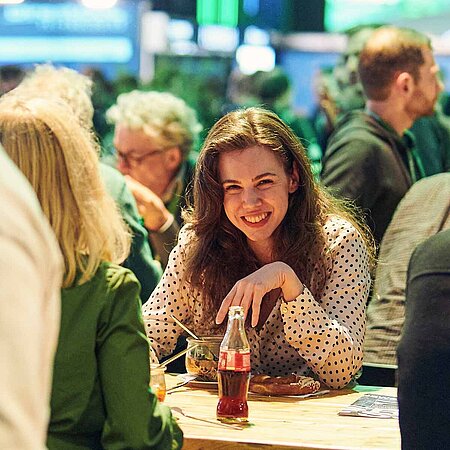 Blick in die Menschengruppe auf der Get-Together. Eine junge Frauch lächelt in die Kamera. Neben ihr befinden sich verschiedene Gruppen im Gespräch. Die Leute trinken und essen dabei.